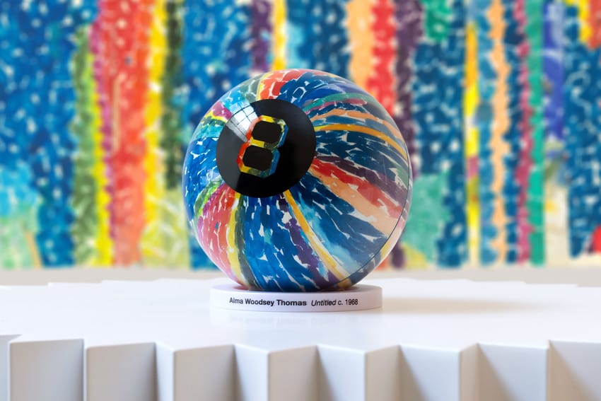 Colorful eight ball sculpture displayed on a white pedestal with vibrant abstract background art.
