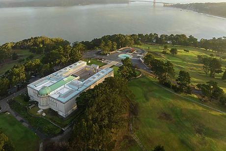 Aerial view of a large museum building surrounded by trees, with a distant bridge and expansive body of water in the background.