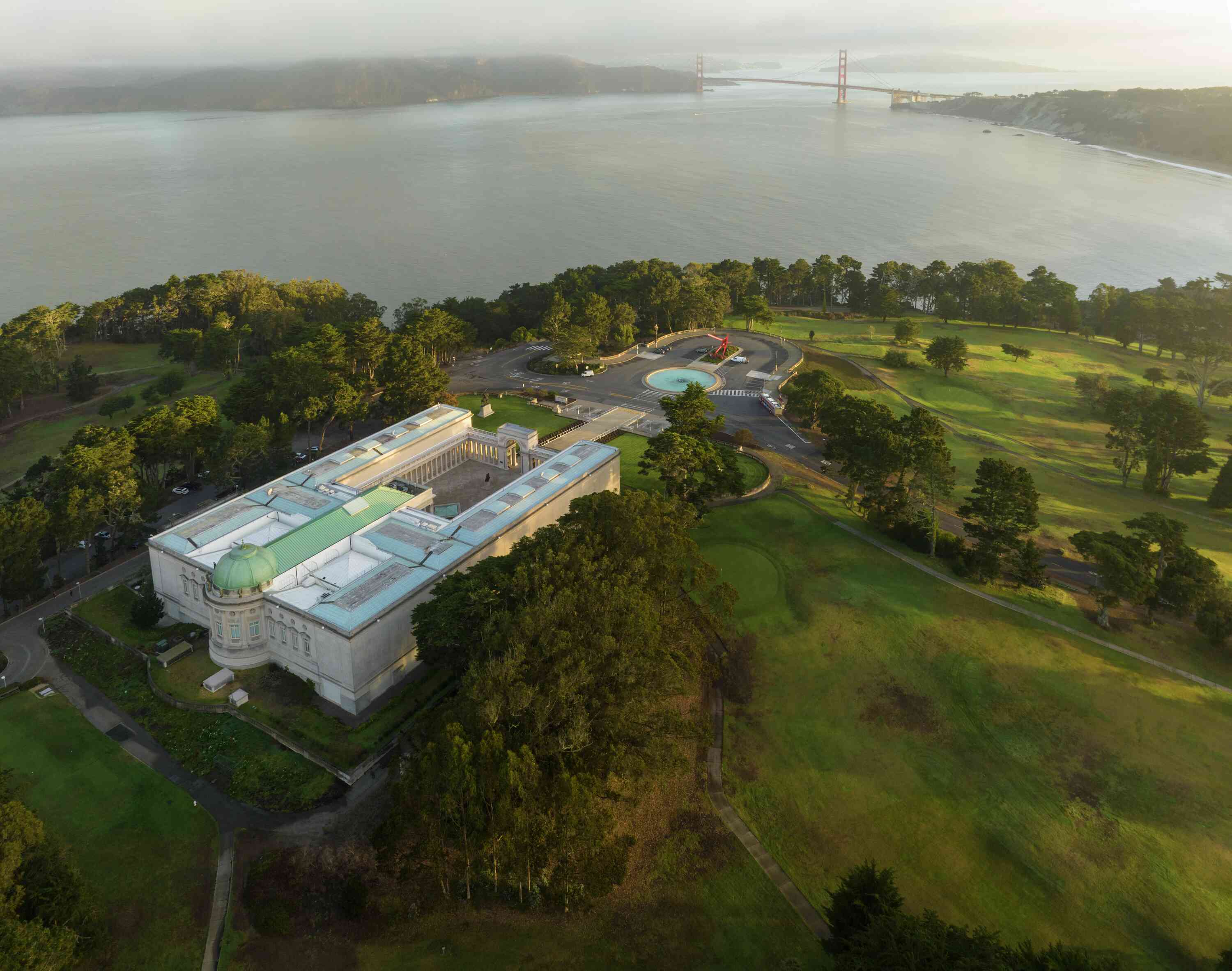 Aerial view of a large museum building surrounded by trees, with a distant bridge and expansive body of water in the background.