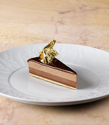 Slice of layered chocolate mousse cake on a white plate, topped with gold leaf, set on a marble surface.