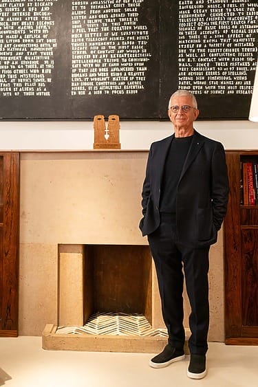 Man in a black suit stands beside a modern fireplace with a decorative art piece and a text-covered board above.