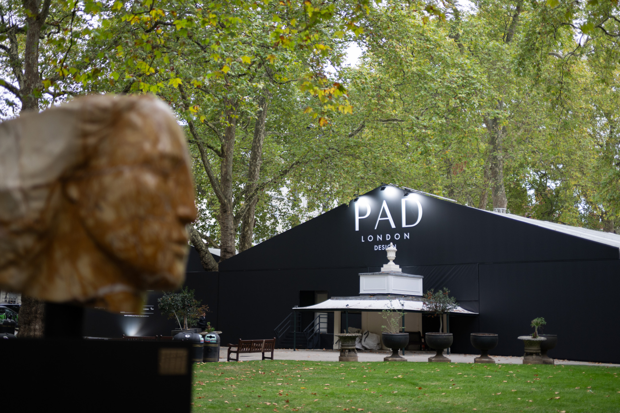 Outdoor view of PAD London design fair entrance, with a blurred sculpture in the foreground and trees in the background.