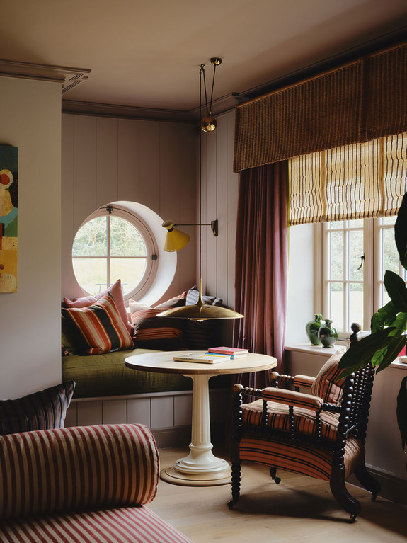 Cozy living room with circular window, striped armchair, sofa, round table, striped curtains, and plants.