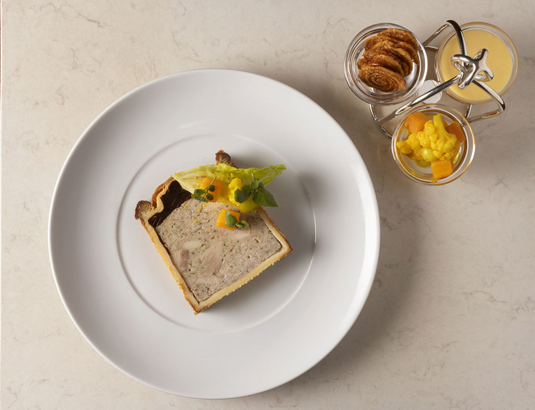 Slice of meat pie on a white plate with vegetable garnish, served alongside jars of mustard and pickled vegetables.