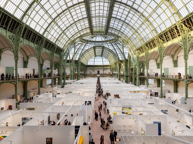 People walking through a large art exhibition in a grand hall with a glass ceiling and green ornate metal framework.