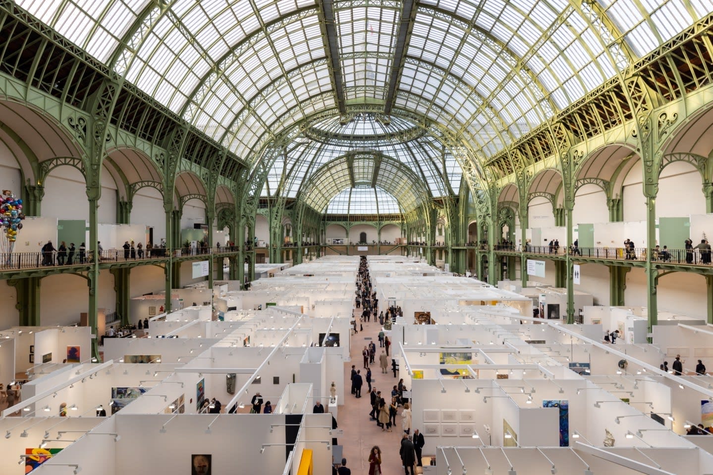 People walking through a large art exhibition in a grand hall with a glass ceiling and green ornate metal framework.