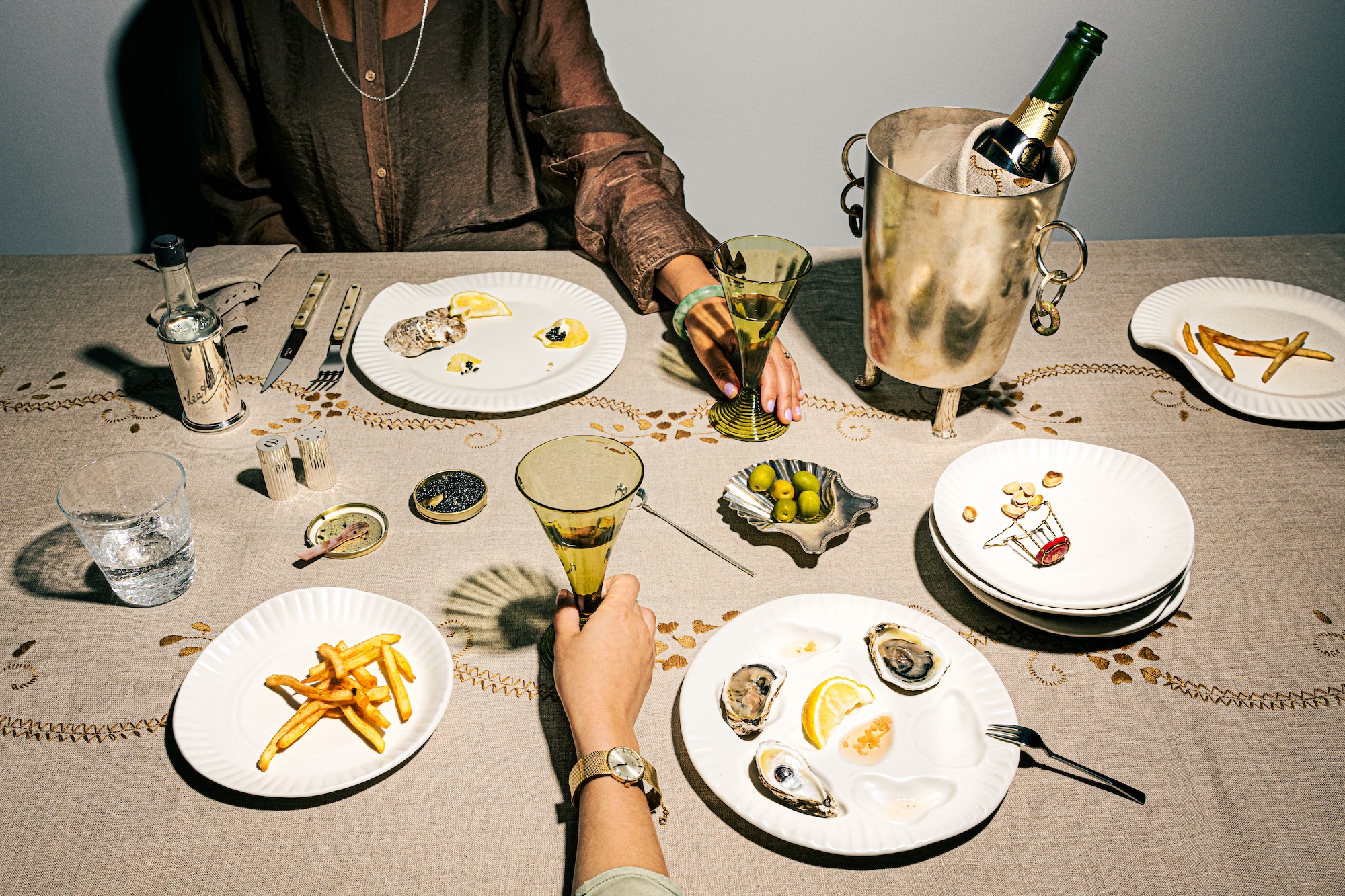 Two people dining with oysters, fries, and champagne on a decorated tablecloth, viewed from above.