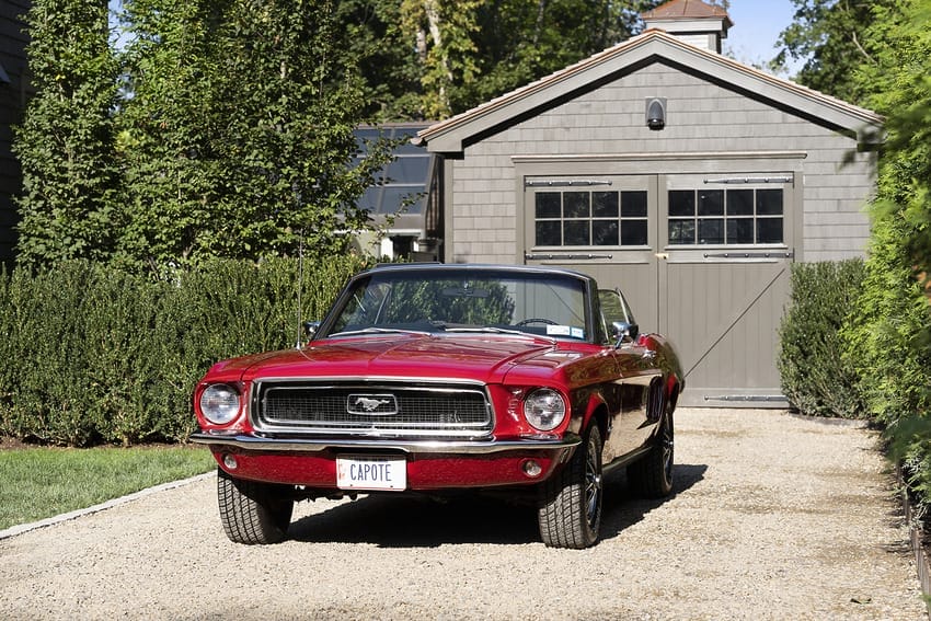 Red vintage car parked in front of a garage with greenery in the background on a sunny day.