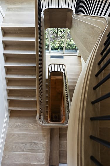 Wooden modern staircase with black railings viewed from above, leading down multiple floors with a window in the background.