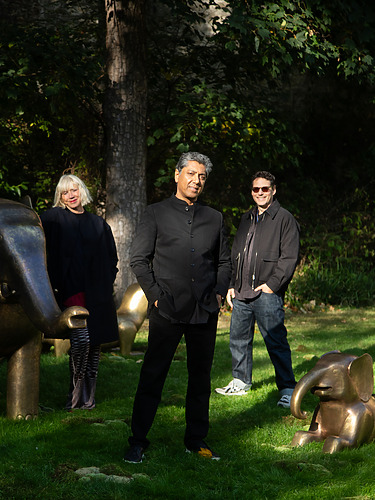 Three people standing in a park with bronze elephant sculptures, surrounded by trees and grass, smiling towards the camera.
