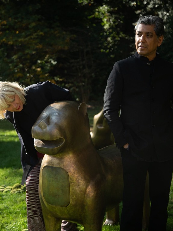 Two people standing beside large bronze lion sculptures in an outdoor setting with greenery in the background.