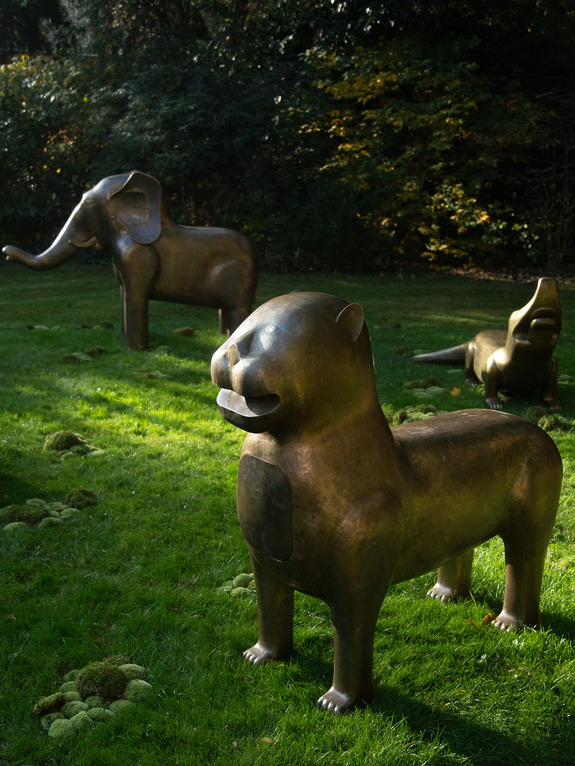 Bronze animal sculptures on grass in a sunlit garden, featuring an elephant, a lion, and a crocodile.