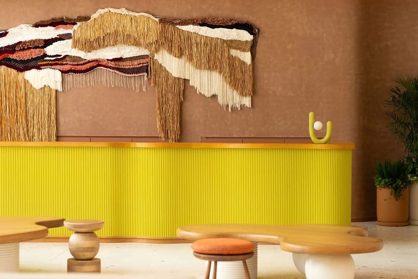 Modern reception area with bright yellow desk, abstract wall art, unique wooden tables, and indoor plants.