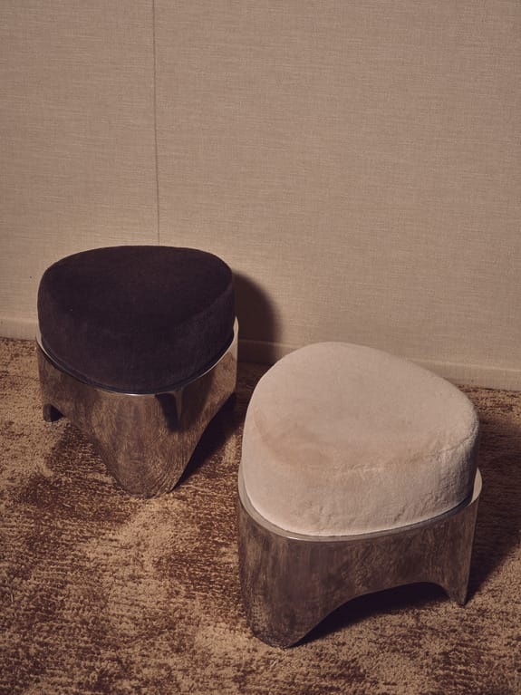 Two modern, cushioned stools with wooden bases on a textured brown carpet against a neutral wall background.