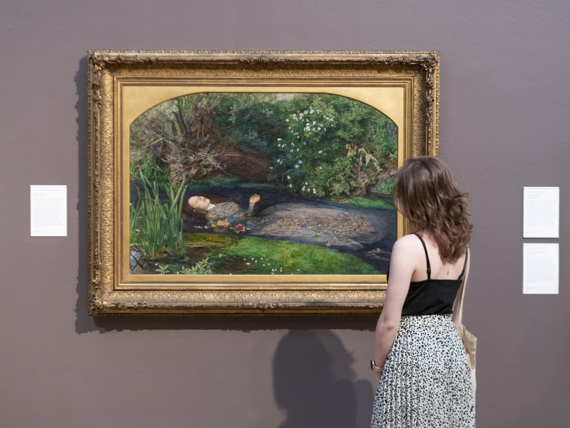 Woman observing a framed painting of a figure laying in a water scene displayed on a gallery wall.