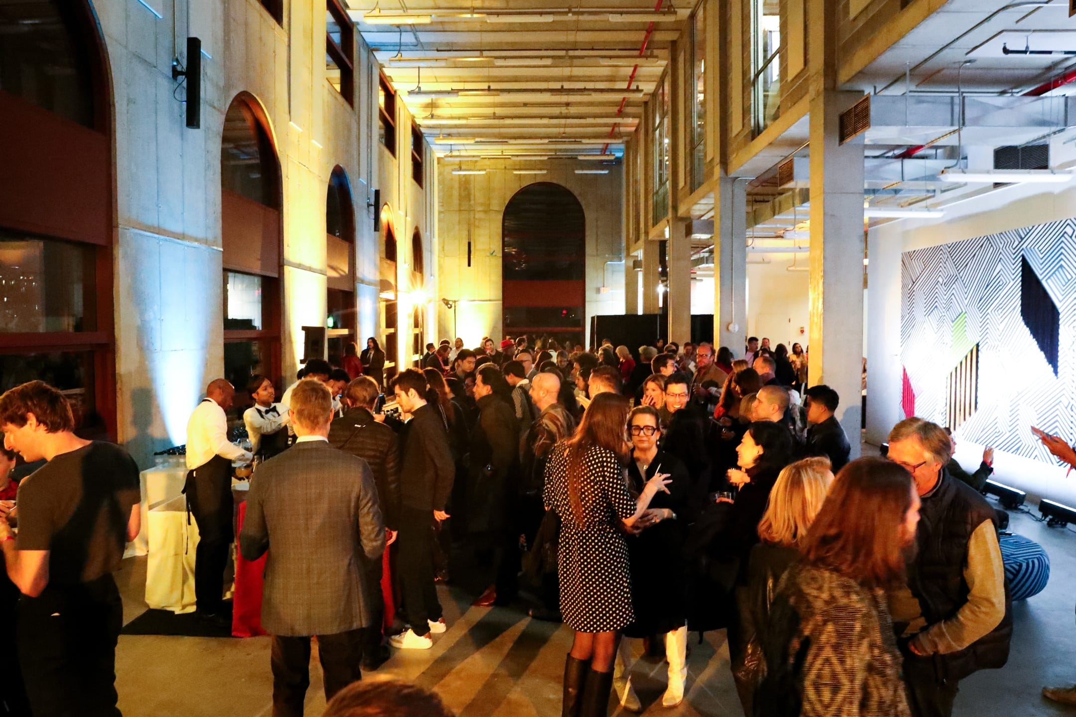 People mingling at a lively indoor social event with modern architecture and wall art in the background.