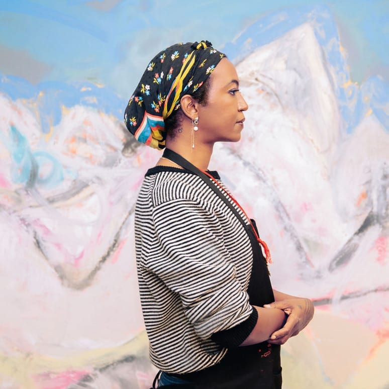 Person standing in front of a colorful abstract mural, wearing a striped top and floral headscarf, viewed in profile.