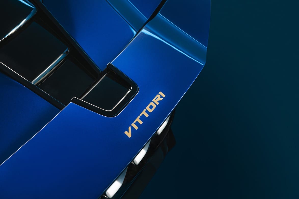 Close-up of a blue sports car hood featuring sleek air vents and the word "VITTORI" in bold gold lettering on the side.