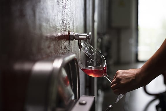 Hand holding a wine glass collecting rosé from a metal spout on a wine tank in a winery setting.