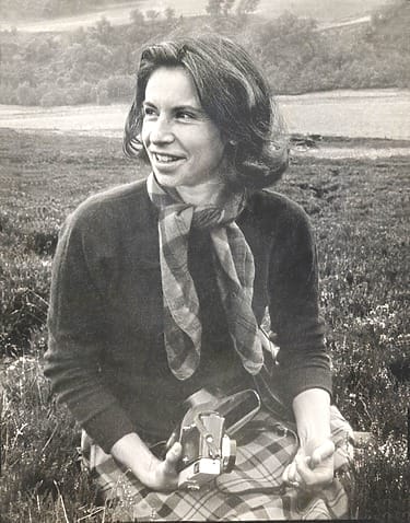Woman sitting on grass in outdoor setting holding a camera, wearing a scarf and plaid skirt, smiling off to the side.