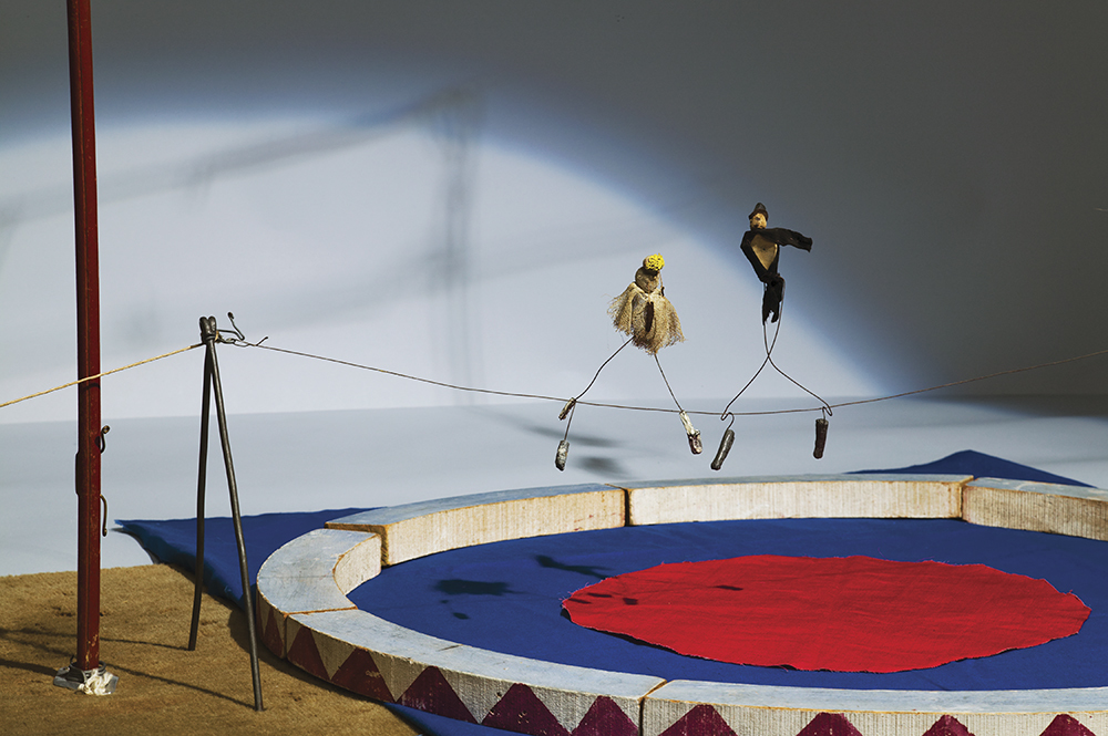 Two figurines on a tightrope above a circus ring with a blue and red mat, set against a shadowy background.