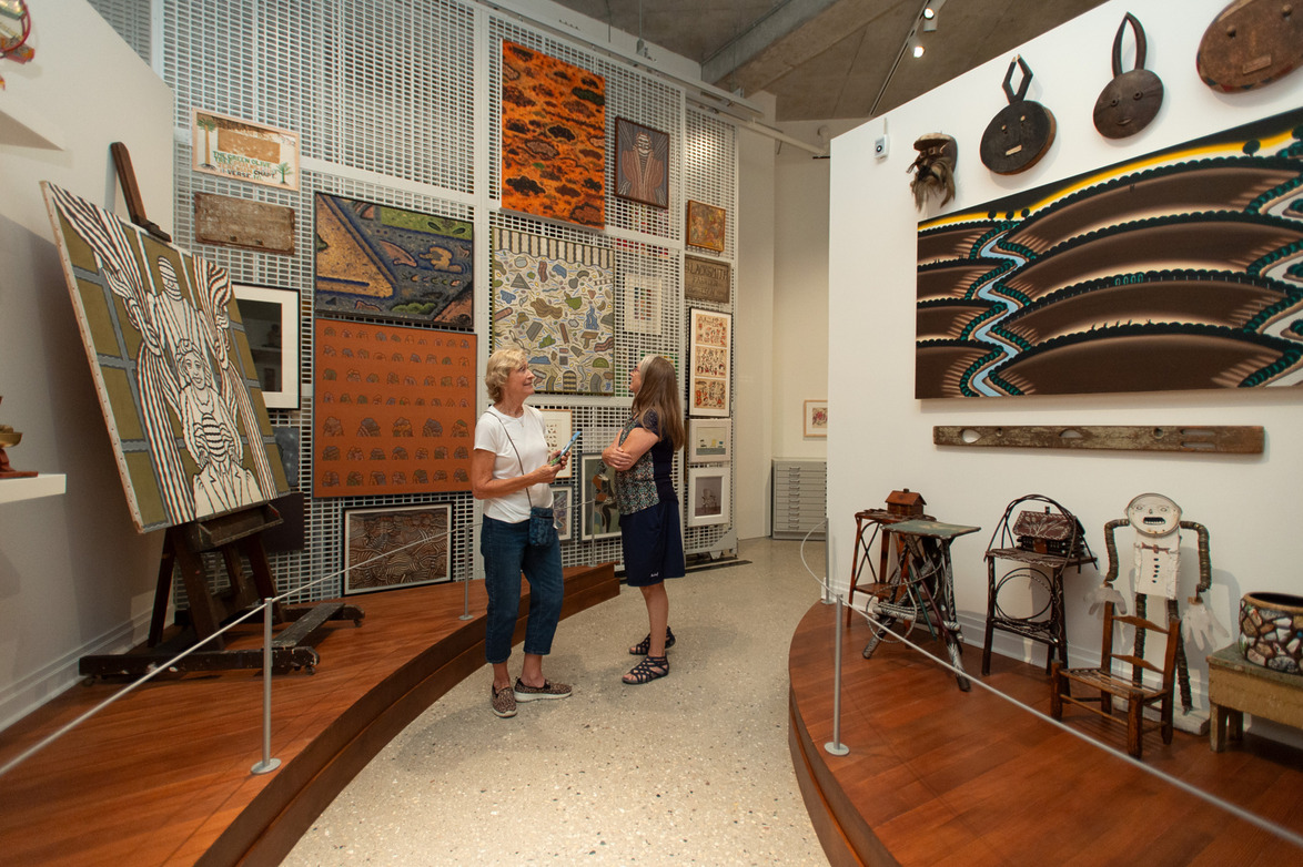 Two people admire Aboriginal art in a gallery, with various paintings and artifacts displayed on the walls and stands.
