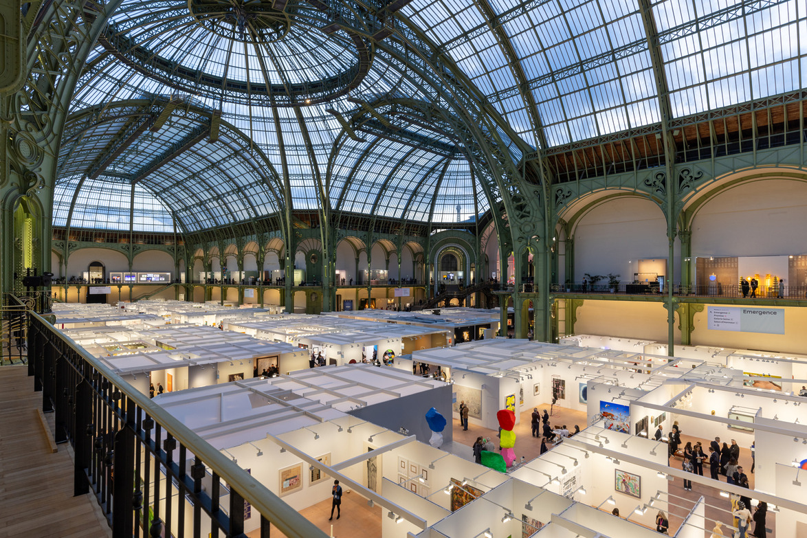 Art exhibition in a large, ornate hall with glass roof and decorative metalwork, showcasing various artworks and visitors.