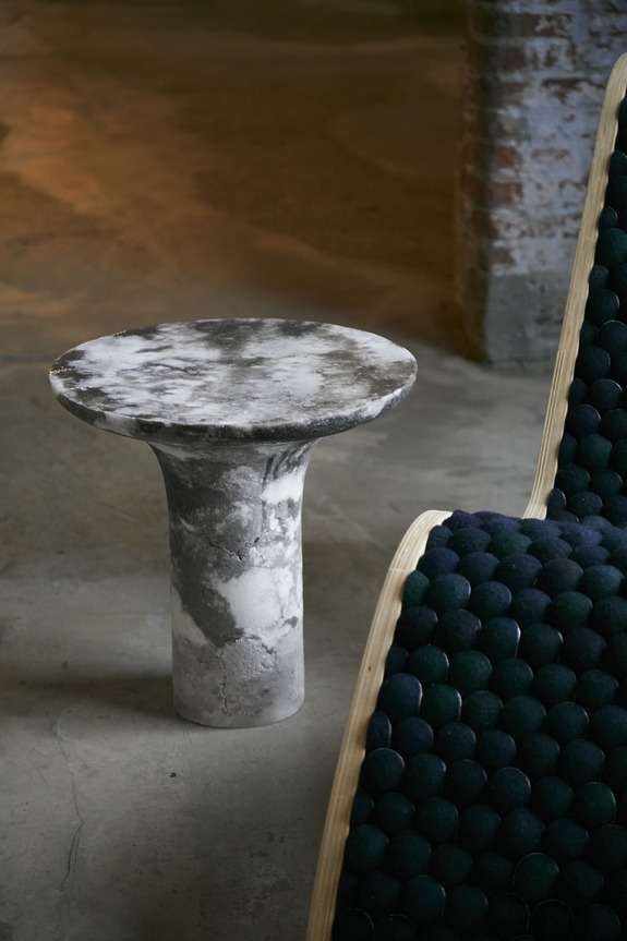 Marble pedestal table next to a textured chair, set on a concrete floor in an industrial-style room.