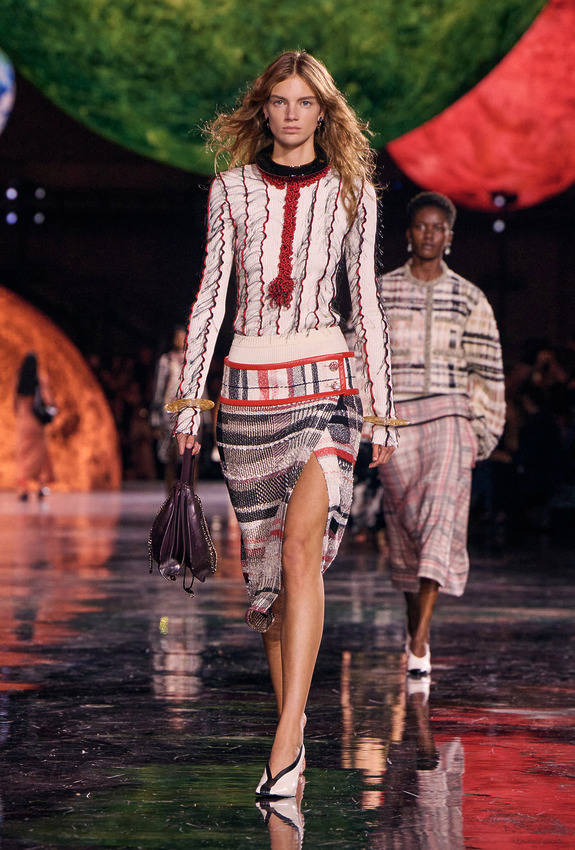 Fashion model in a stylish knit ensemble walking on a reflective runway with vibrant spherical backdrops.