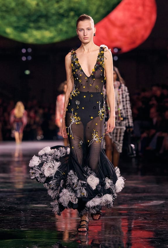 Model walking on a fashion runway wearing a stylish black sheer gown adorned with floral and ruffled details.