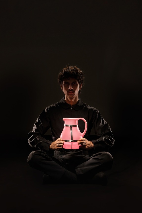 Person sitting cross-legged in the dark holding a glowing pink pitcher, wearing a black outfit.
