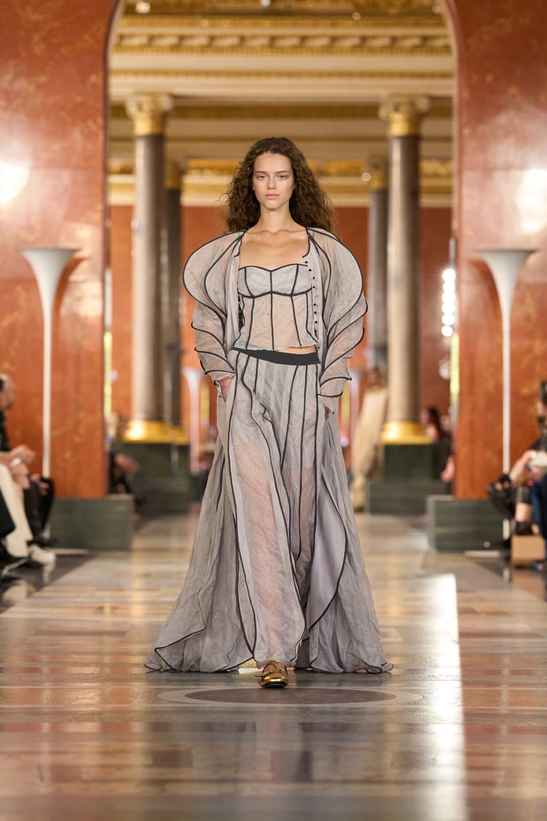 Model walking on runway in elegant sheer gray dress with architectural columns in background at fashion show.