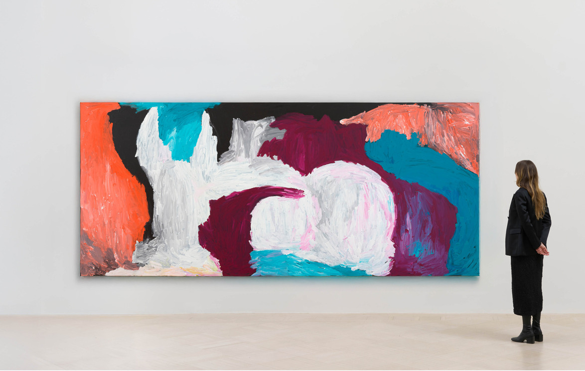 A person observing a large colorful abstract painting with vibrant hues of orange, pink, blue, and white in a gallery setting
