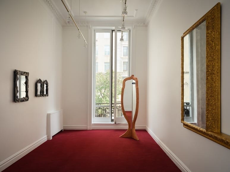 Minimalist room with red carpet, large window, ornate mirror, and unique freestanding mirror on a wooden stand.