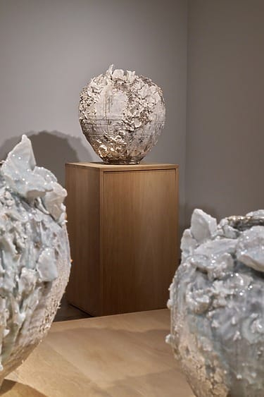 Ceramic sculpture with textured, rock-like surface on a wooden pedestal in an art gallery setting.
