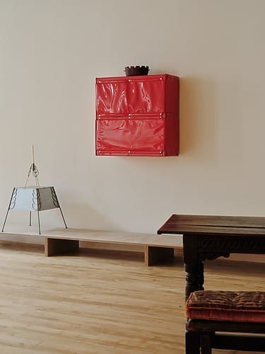 Red wall-mounted cabinet above wooden bench and table in a minimalist room with light wooden floor.