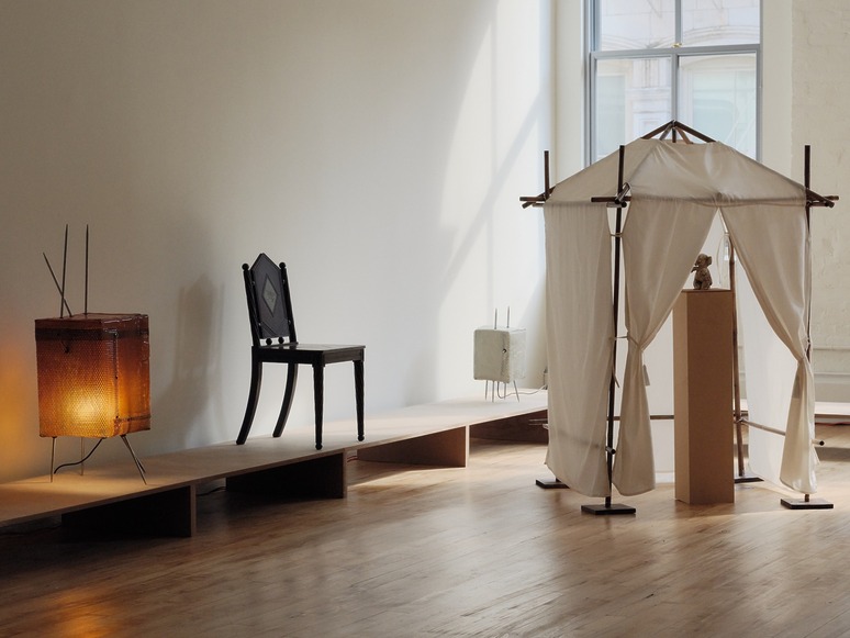 Art installation with wooden platform, chair, illuminated box, and fabric structure in a sunlit gallery space.