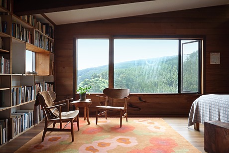 Cozy room with wooden walls, bookshelves, and chairs, featuring a large window with a scenic mountain view.