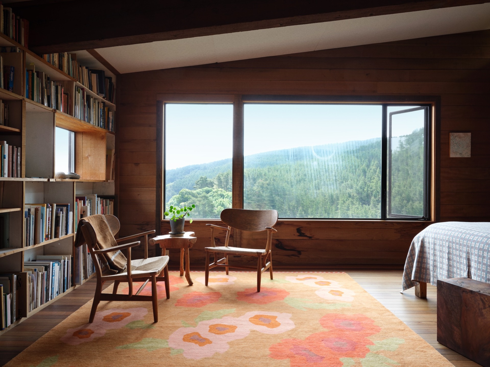 Cozy room with wooden walls, bookshelves, and chairs, featuring a large window with a scenic mountain view.