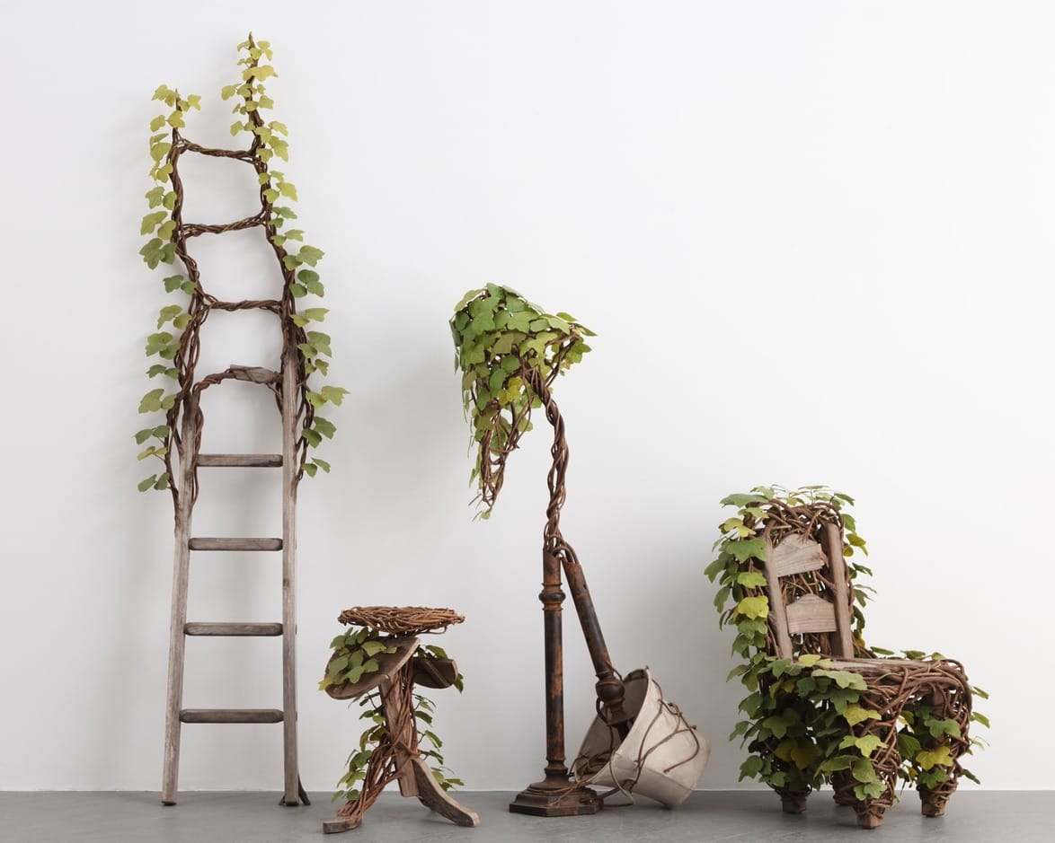 Wooden ladder, stools, lamp, and chair wrapped with green leaves against a white wall, showcasing nature-inspired design.