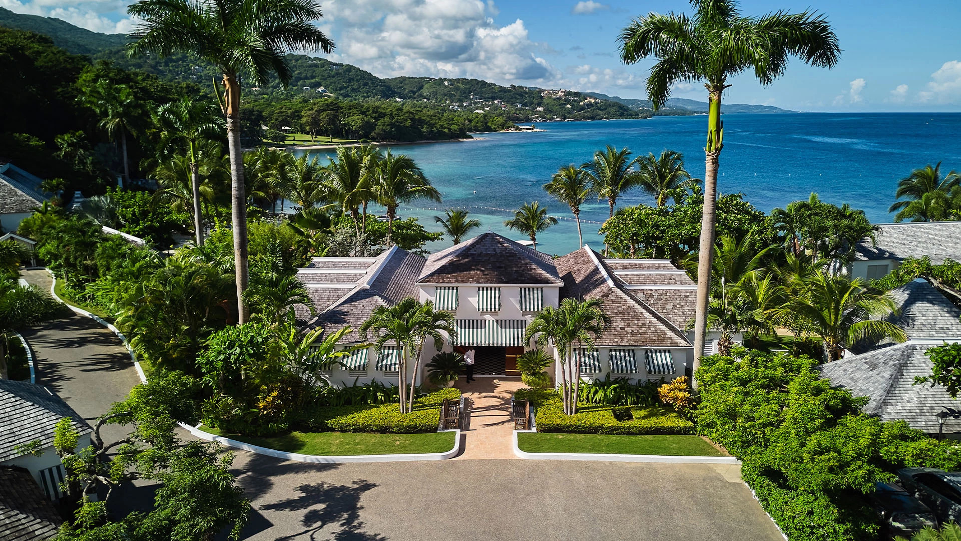 Tropical beachfront villa with palm trees, lush gardens, and ocean view under a sunny sky.
