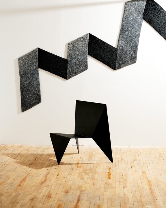 Abstract black chair on wooden floor with zigzag black and gray wall hanging in a bright, minimalist gallery space.