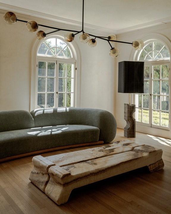 Modern living room with a curved sofa, unique stone coffee table, large windows, and a contemporary light fixture.