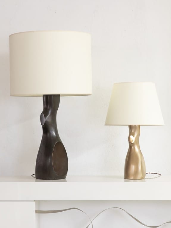 Two modern table lamps, one dark and tall, the other short and gold, placed on a white shelf against a plain wall.