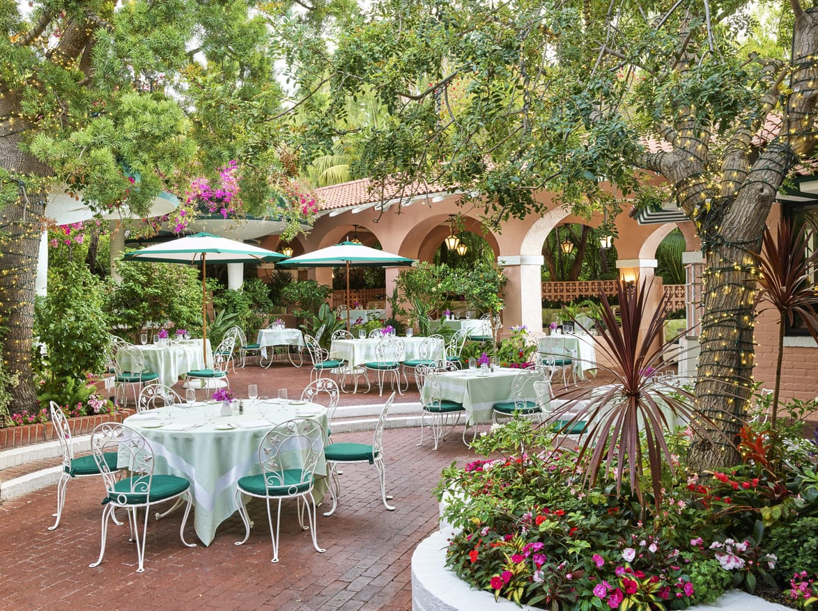 Outdoor restaurant patio with tables and chairs surrounded by green trees and colorful flowers in a sunny garden setting.