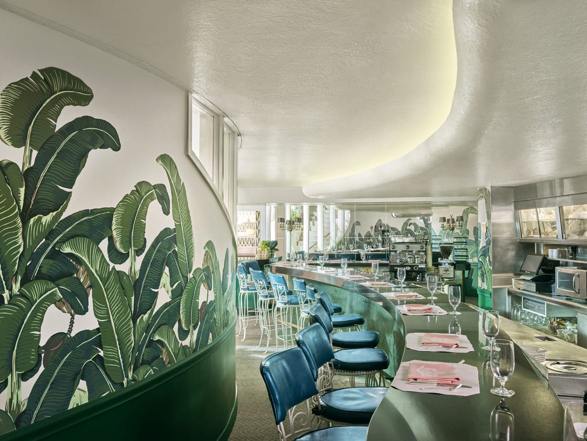 Retro diner interior with curved counter, blue stools, tropical wall mural, and set tables with wine glasses and napkins.