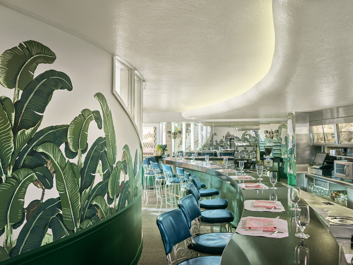 Retro diner interior with curved counter, blue stools, tropical wall mural, and set tables with wine glasses and napkins.