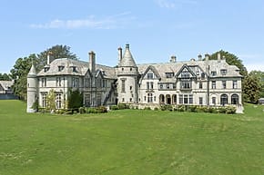 Large historic mansion with stone facade, numerous chimneys, and lush green lawn on a clear, sunny day.