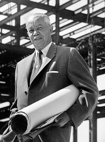 Architect holding blueprints, standing in front of a construction site with steel framework in the background.