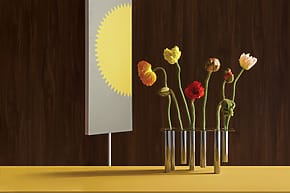 Modern art display with vibrant flowers in glass vases on a yellow table and a yellow sun artwork on a dark wall.